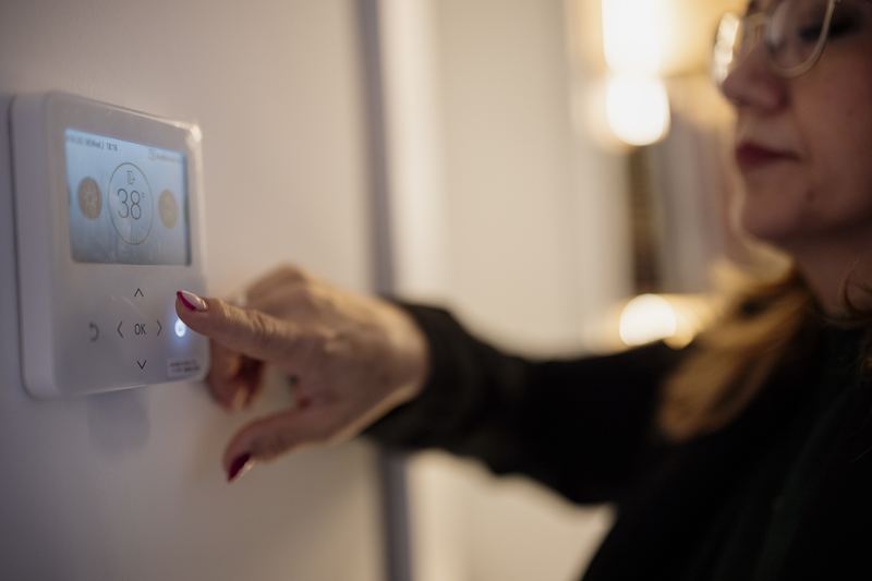 A woman is adjusting a digital thermostat mounted on a wall. She looks focused on setting the temperature. This appears to take place in a home setting.